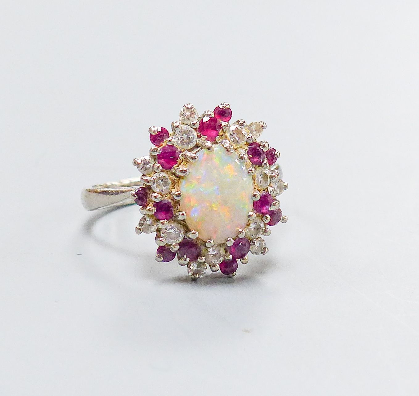 An 18ct white metal, white opal, ruby and diamond set oval cluster ring, size N, gross weight 6.1 grams.